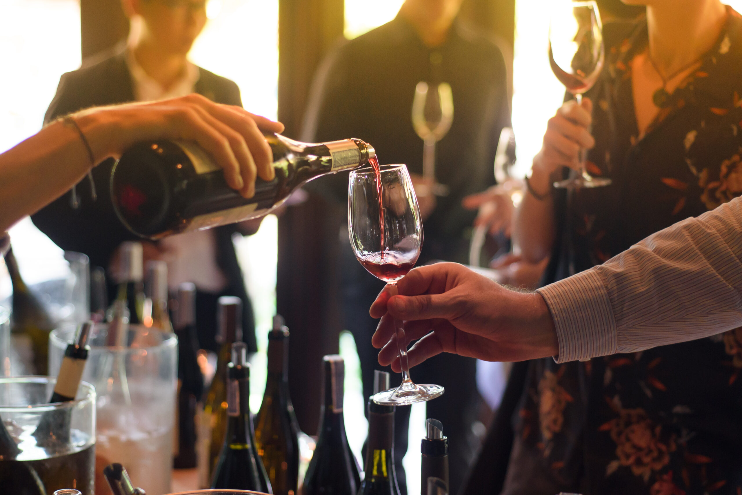 A sommelier pours red wine into a glass at a lively wine-tasting event, with elegantly dressed guests in the background holding wine glasses and engaging in conversation.