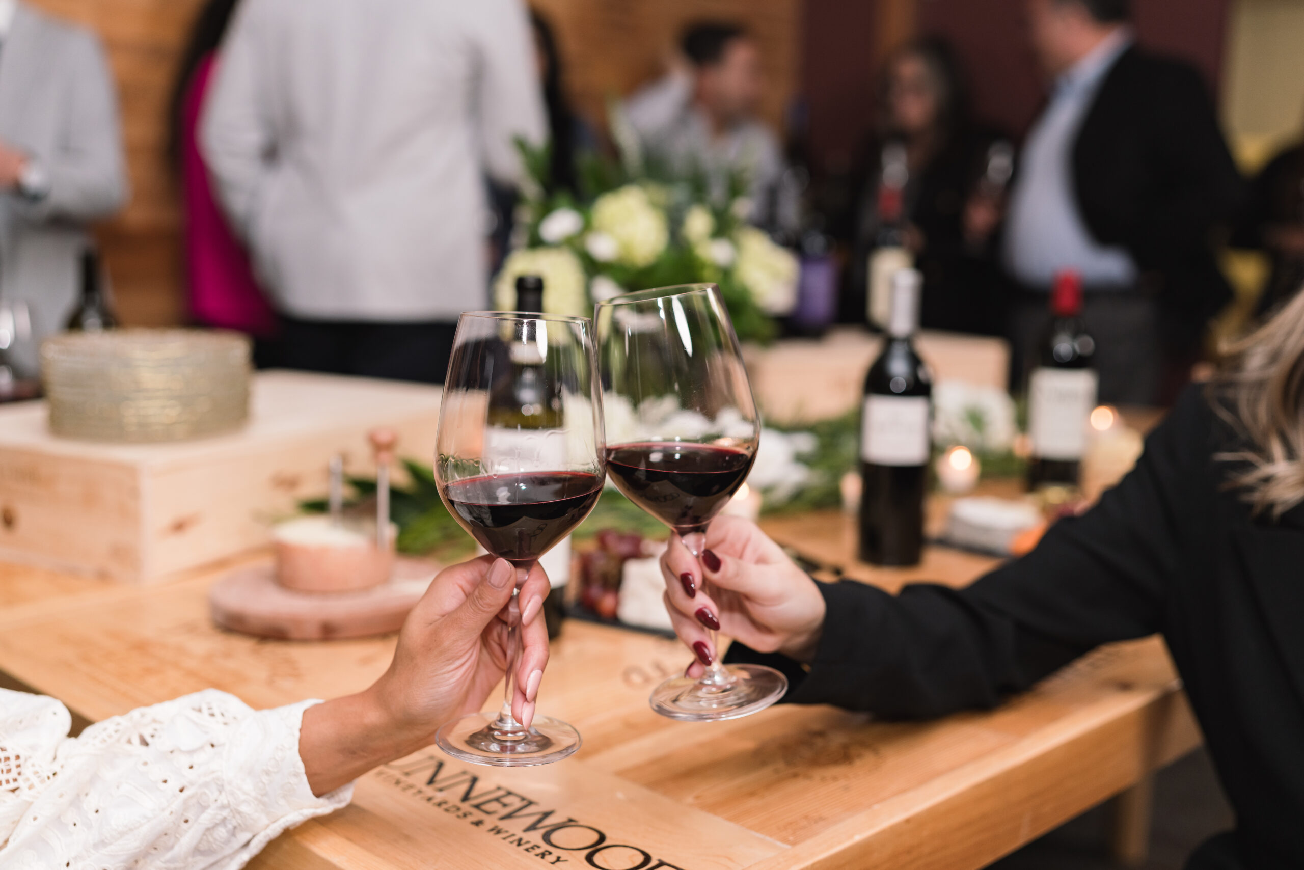 Two elegantly dressed guests toast with glasses of red wine at a sophisticated wine tasting event, with a beautifully decorated table and blurred background of mingling attendees.