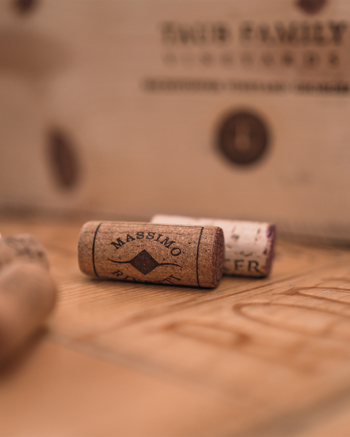 Close-up of a Massimo wine cork resting on a wooden table, with another cork and a blurred wine crate in the background, evoking a rustic and elegant wine-tasting atmosphere.
